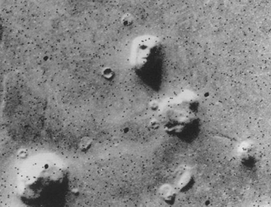 "Face On Mars" photograph taken by the Viking 1 orbiter and released by NASA/JPL on July 25, 1976. The large mesa sculptured as a face is 1.2 miles (2 km) long in Cydonia Mensae, at 40.75° north latitude and 9.46° west longitude. When the image was originally acquired, Viking chief scientist Gerry Soffen dismissed the “Face on Mars” in image 035A72 as a “trick of light and shadow.” However, other computer analysts and plasma physicist John E. Brandenburg, Ph.D., say the 5-sided pyramid and other structures not far from the sculpted face (below) are evidence of an ancient civilization in Cydonia Mensae and Galaxias Chaos to the east where two atmospheric hydrogen bombs left trinitite, xenon-129, Argon 40 and krypton evidence that is now being presented at serious science conferences by Dr. Brandenburg. The dark spots all over this original 1976 Viking 1 images are attributed to “static digital dropouts.”