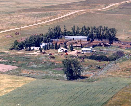 Farm house has slanted roof at center. Long gray building north of house is barn. Left of barn is pig corral fenced off by sheet metal walls. One half mile east (off right) is water pond. Aerial image © by Torrey and Myra Briese.