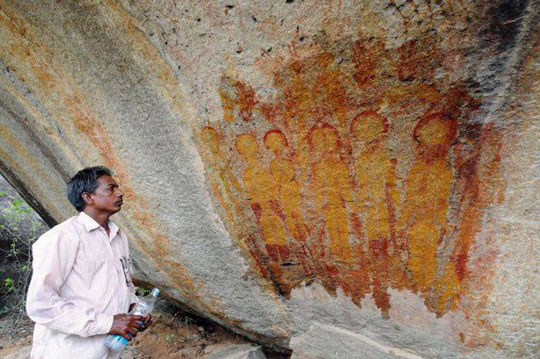 Archaeologist J. R. Bhagat standing next to one of the cave rock paintings of gold-robed beings with large, rounded heads.